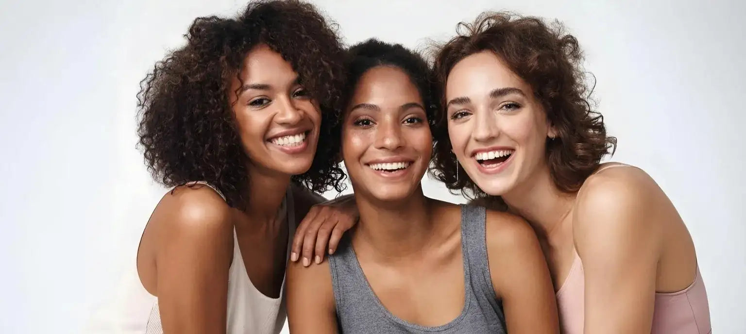 Three smiling women standing close together against a light background in a lifestyle beauty portrait